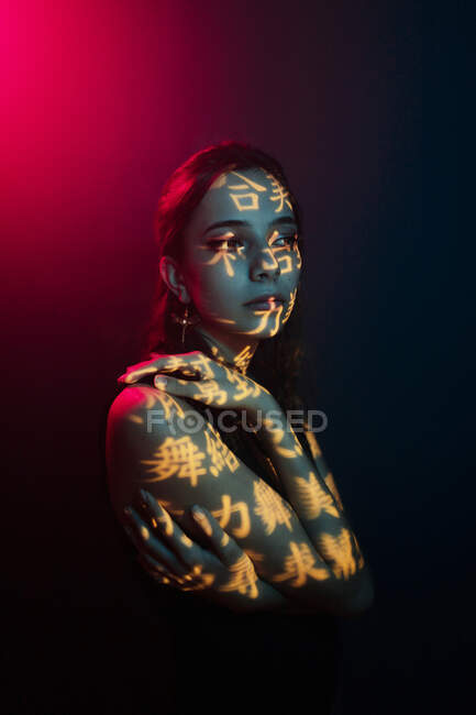 Fashionable young female model with light projection in shape of oriental hieroglyphs looking away in dark studio with red illumination — Stock Photo