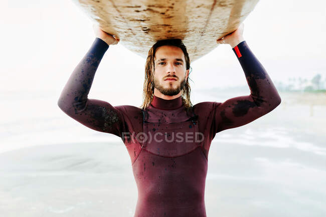 Portrait of young surfer man with long hair and beard dressed in wetsuit standing looking at camera on the beach with the surfboard above head during sunrise — Stock Photo