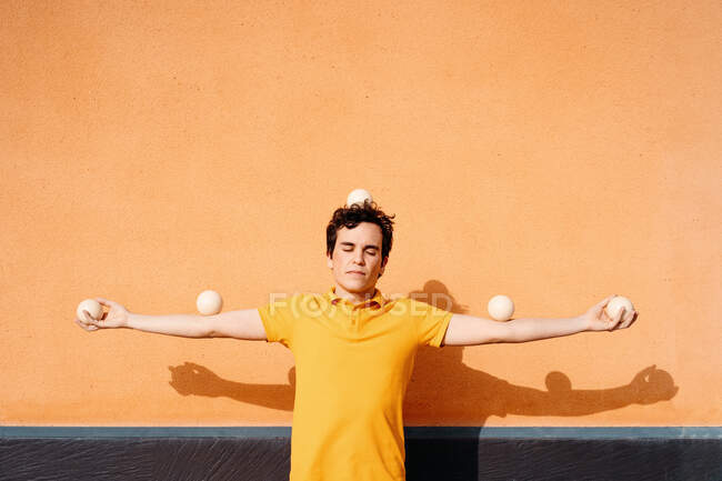 Спокойный неподвижный молодой мужчина в ярко-желтой рубашке с жонглирующими шарами на голове и вытянутыми руками, стоящими с закрытыми глазами у оранжевой стены — стоковое фото