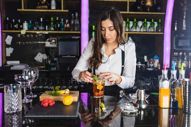 Young female barkeeper in stylish outfit preparing cocktail with lemon slices while standing at counter in modern bar — Stock Photo