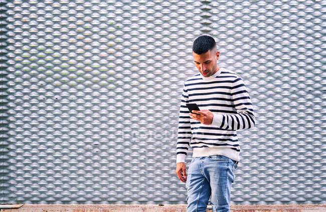 Homme hispanique adulte en vêtements décontractés debout près du mur ornemental gris et la navigation sur les médias sociaux sur smartphone sur la rue de la ville — Photo de stock