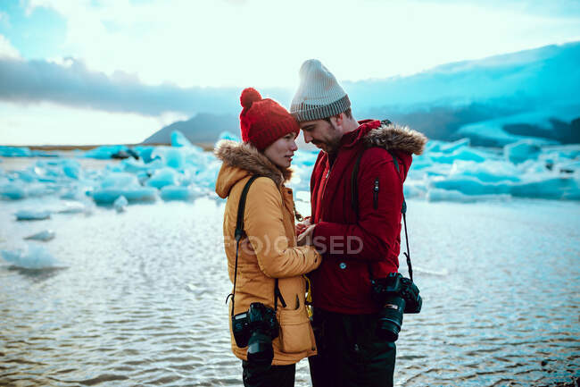Vista lateral do homem e da mulher novos no desgaste do inverno com câmeras profissionais que estão na costa perto da água com gelo — Fotografia de Stock