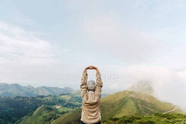 Visão traseira de viajante sênior irreconhecível levantando e esticando os braços de manhã em terreno montanhoso — Fotografia de Stock