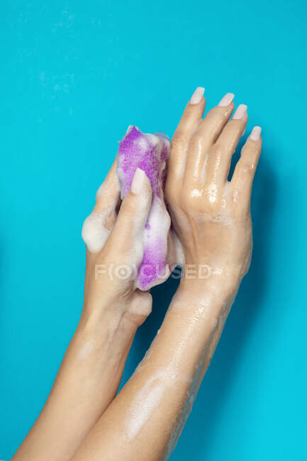 Mano di raccolto anonimo femminile spugna bagno spremitura con schiuma bianca contro sfondo blu — Foto stock