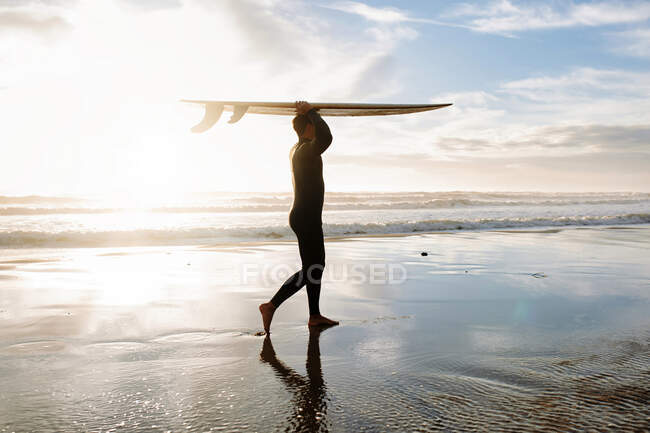 Вид сбоку человека-серфера, одетого в гидрокостюм, идущего по пляжу с доской для серфинга над головой утром с восходом солнца на заднем плане — стоковое фото