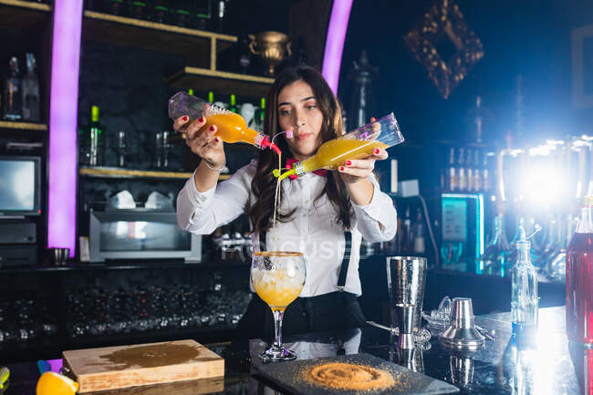 Focused female barkeeper in stylish outfit adding yellow colorant liquid from bottle into glass while preparing cocktail standing at counter in modern bar — Stock Photo