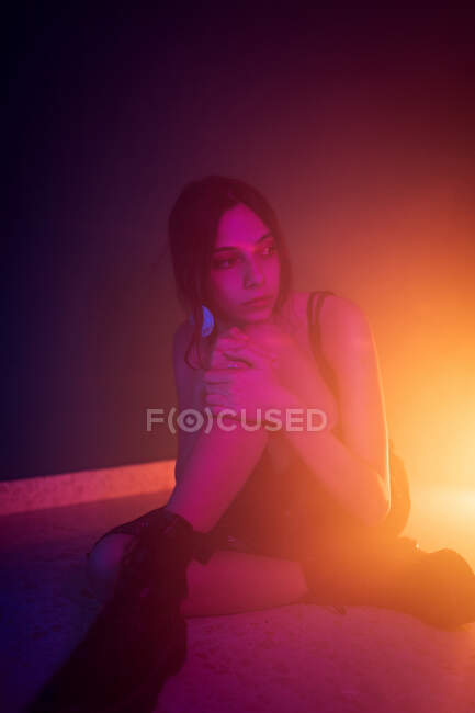 Tranquil modelo femenina joven en vestido sentado en el suelo y apoyado en la rodilla mientras mira hacia otro lado en el estudio oscuro con luces de colores - foto de stock