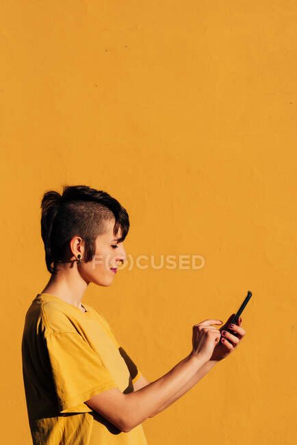Сторона зору сучасної жінки з стильною зачіскою і пірсингом з використанням смартфона для текстового повідомлення в соціальних мережах на жовтому тлі. — стокове фото