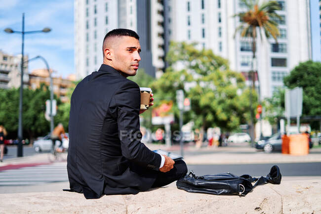 Low angle side view of serious pensive young Hispanic man in formal black suit looking away thoughtfully while sitting with digital gadget in hand on urban street — Stock Photo