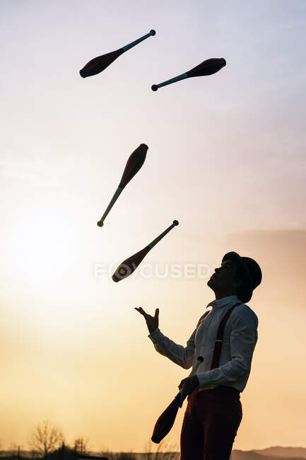 Visão lateral de baixo ângulo de artista de circo masculino irreconhecível em clubes de malabarismo chapéu contra o céu por do sol — Fotografia de Stock