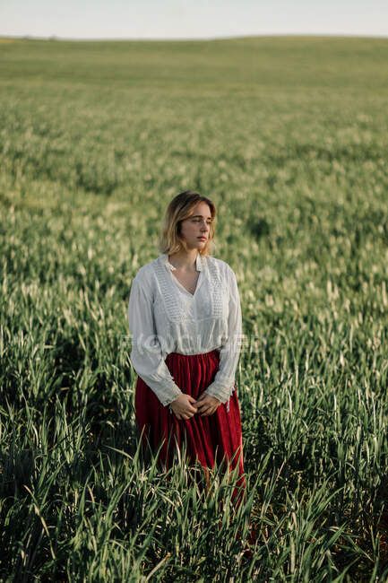Calm young female dressed in old fashioned blouse and skirt standing alone among tall green grass in cloudy summer day in countryside — Stock Photo