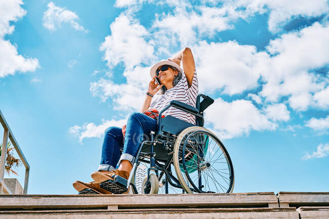 Mature disabled female sitting in wheelchair and talking on mobile phone near stairway against blue sky in city — Stock Photo