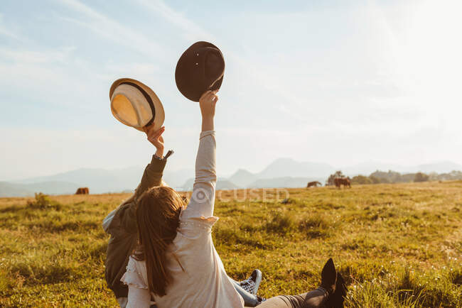 Visão traseira de amigos femininos irreconhecíveis levantando as mãos com chapéus sentados no gramado à luz do sol e desfrutando de liberdade durante as férias de verão — Fotografia de Stock