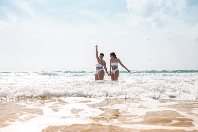 From below cheerful female friends holding hands in swimsuits in foamy ocean near sandy beach under blue cloudy sky in sunny day — Stock Photo