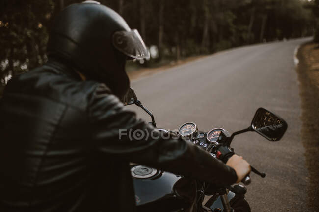 Back view of male in leather jacket and safety helmet driving on motorcycle on asphalt route between forest — Stock Photo