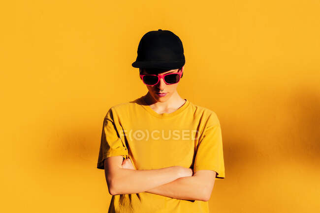 Mujer joven con gorra y camiseta estilo calle con los brazos cruzados de pie contra la pared amarilla en la calle de la ciudad - foto de stock