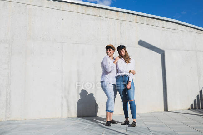 Повне тіло молодих позитивних друзів-жінки в модних вбраннях і капелюхах, що стоять на прогулянці біля сірої стіни в сонячний день під блакитним небом — стокове фото