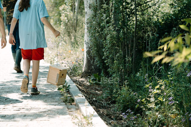 Visão traseira da adolescente irreconhecível carregando caixa de nidificação de madeira artesanal para pássaros enquanto se reúne com voluntários durante a campanha ambiental no parque de verão — Fotografia de Stock
