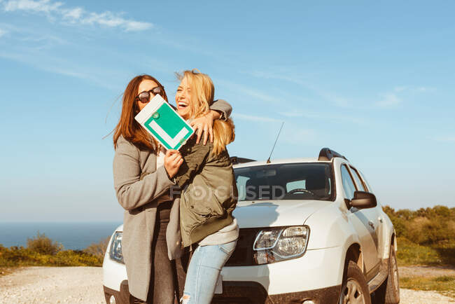 Alegre joven novias con señal de marcha baja de pie en el coche blanco en la orilla del mar y abrazar tener viaje - foto de stock