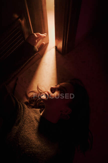 Сверху тихая молодая женщина лежит на полу в темной комнате со светящимся от открытой двери светом, глядя в сторону — стоковое фото