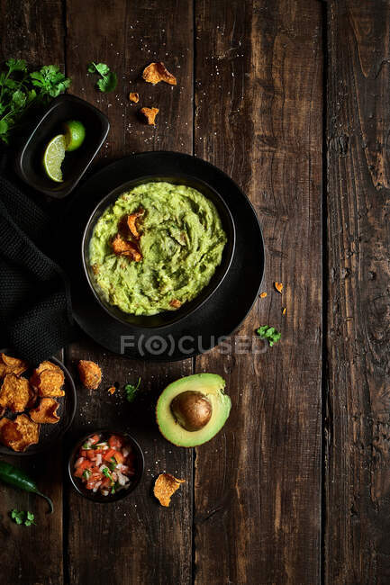 Вид сверху на чашу свежего гуакамоле, расположенную рядом с авокадо, и чипсы на мокрой столешнице — стоковое фото