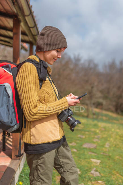 Vista lateral del explorador femenino alegre con la mochila de pie en la naturaleza cerca de la casa y el teléfono móvil de navegación - foto de stock