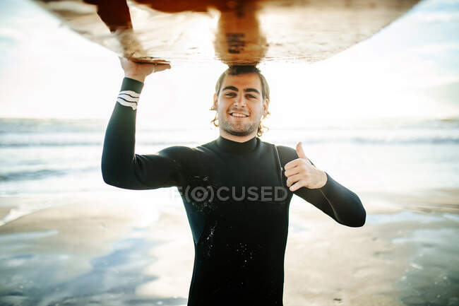 Портрет молодого щасливого серфінгу, одягненого в гідрокостюм, стоїть з великими пальцями вгору, дивлячись на камеру на пляжі з дошкою для серфінгу над головою під час сходу сонця — стокове фото