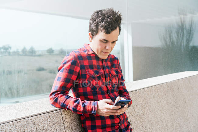 Молодий чоловік у пробній сорочці переглядає мобільний телефон поблизу сучасного будинку зі скляними стінами та кам 