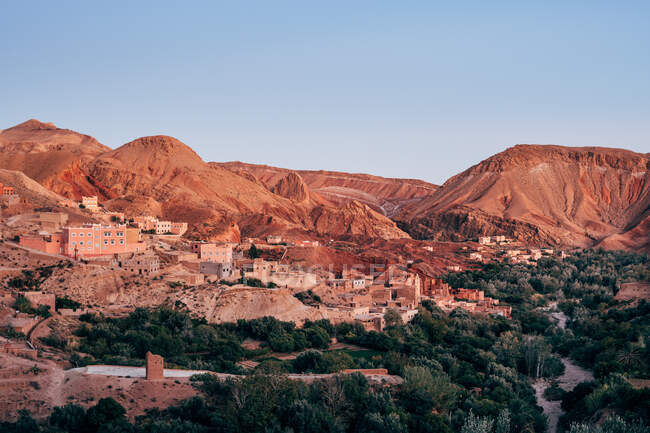 D'en haut de vieux bâtiments en pierre sur la pente de grandes montagnes colorées parmi les plantes vertes avec un ciel bleu clair sur le fond au Maroc — Photo de stock