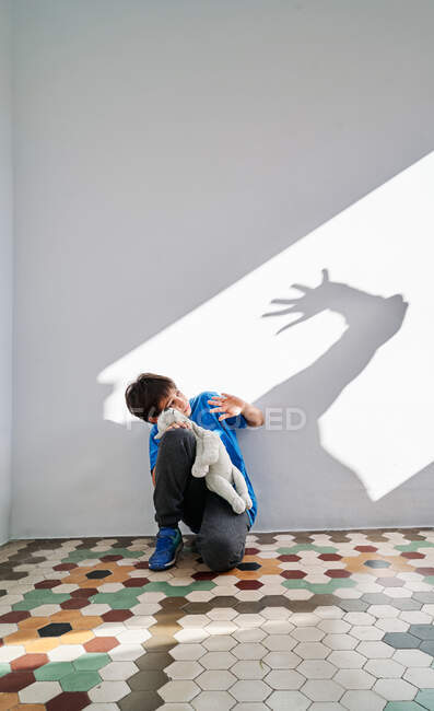 Niño asustado con juguete en las manos sentado cerca de la pared con sombra de padre enojado violento con el brazo levantado - foto de stock