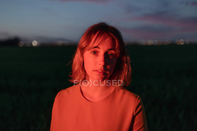 Мрійлива молода жінка в вінтажному стилі одягається, дивлячись на камеру вдумливо, стоячи на самоті в темному трав'яному полі проти хмарного сонцестояння в сутінках — стокове фото