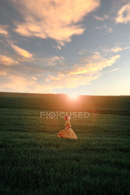 Young female in vintage style looking away thoughtfully while walking alone in grassy field at sunset time in summer evening in countryside — Stock Photo