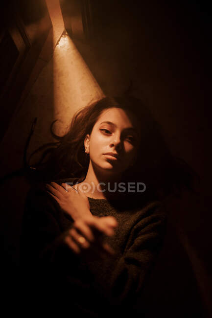 From above of silent young female lying on floor in dark room with light glowing from opened door looking away — Stock Photo