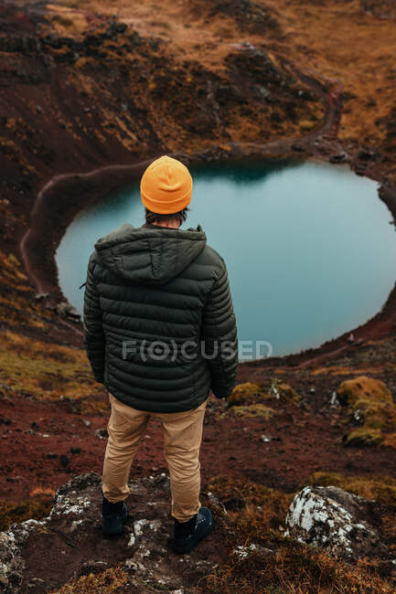 Back view of young tourist in winter wear looking at pound and mountain river between stone hill — Stock Photo