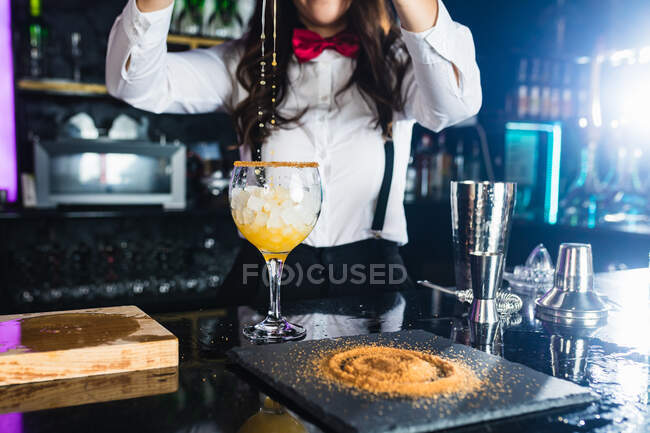 Cropped unrecognizable female barkeeper in stylish outfit adding yellow colorant liquid from bottle into glass while preparing cocktail standing at counter in modern bar — Stock Photo