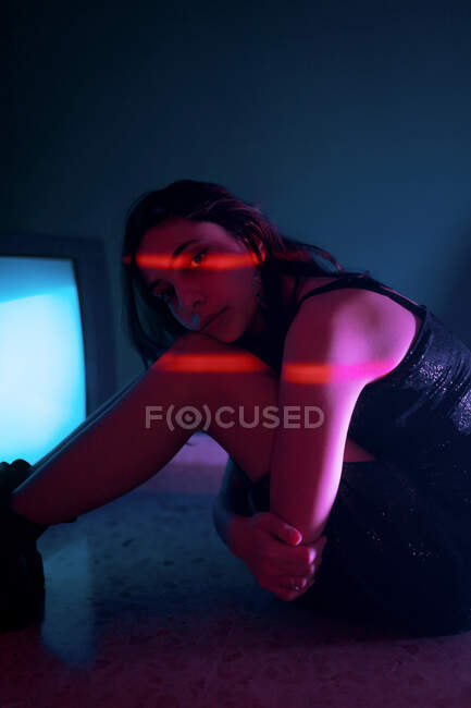 Vista lateral do modelo feminino em vestido preto sentado olhando para a câmera no chão perto de brilhar televisão antiga no estúdio escuro — Fotografia de Stock