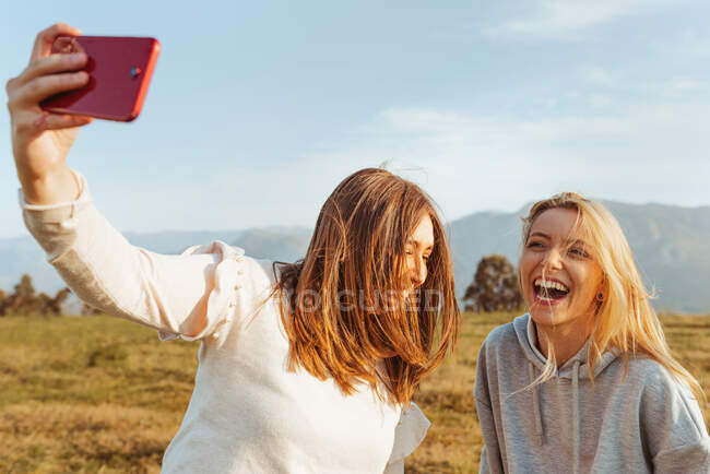 Cheerful young girlfriends taking self photo with smartphone in countryside of mountains — Stock Photo