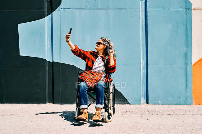 Corpo inteiro vista lateral de independente deficiente maduro fêmea tomando selfie no celular perto de parede pintada na rua — Fotografia de Stock