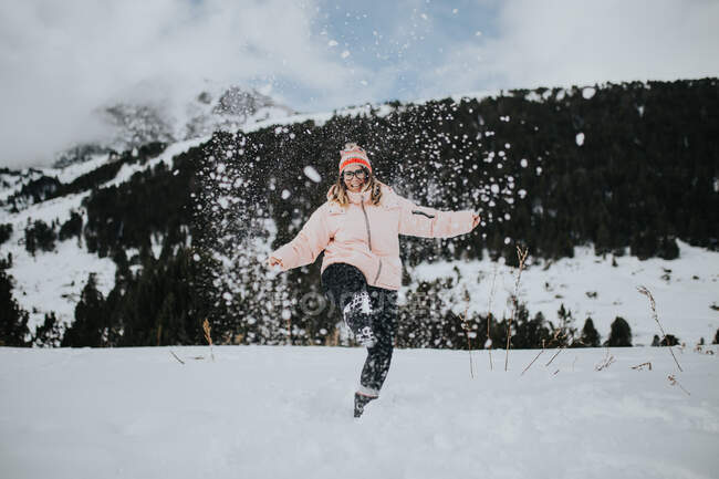 Junger fröhlicher Tourist mit Brille und hochgestrecktem Bein blickt in die Kamera und amüsiert sich zwischen Feld im Schnee in der Nähe des Berges — Stockfoto
