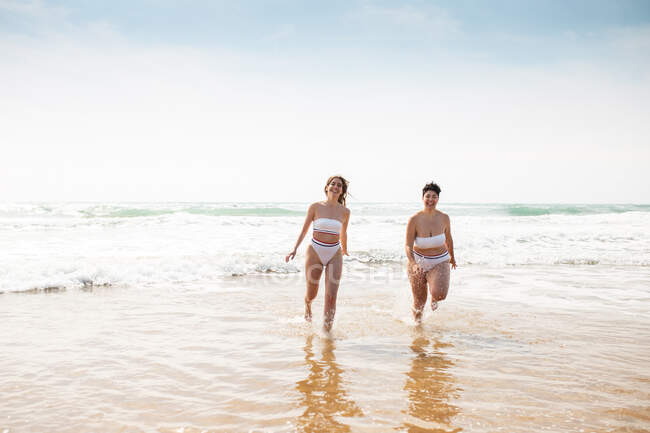 Allegre amiche in costume da bagno in mare schiumoso vicino alla spiaggia di sabbia sotto il cielo nuvoloso blu nella giornata di sole — Foto stock