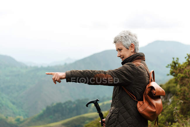 Side view of elderly woman with backpack and trekking stick standing on grassy slope towards mountain peak during trip in nature pointing with finger — Stock Photo