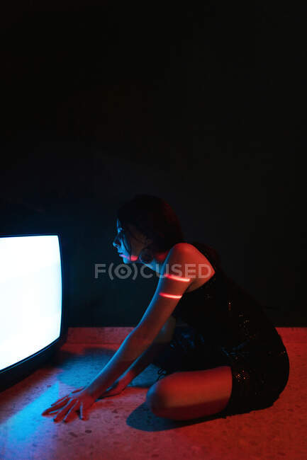 Vista lateral do modelo feminino em vestido preto sentado no chão perto de uma televisão antiga brilhante no estúdio escuro — Fotografia de Stock