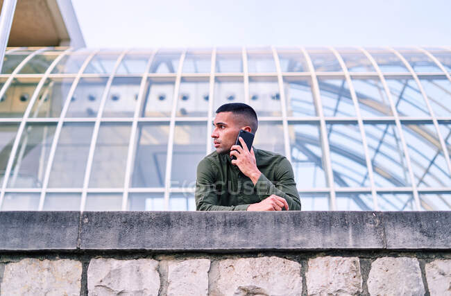 Baixo ângulo do homem hispânico adulto na camisa inclinada na parede de pedra e falando no celular fora do edifício de vidro contemporâneo — Fotografia de Stock