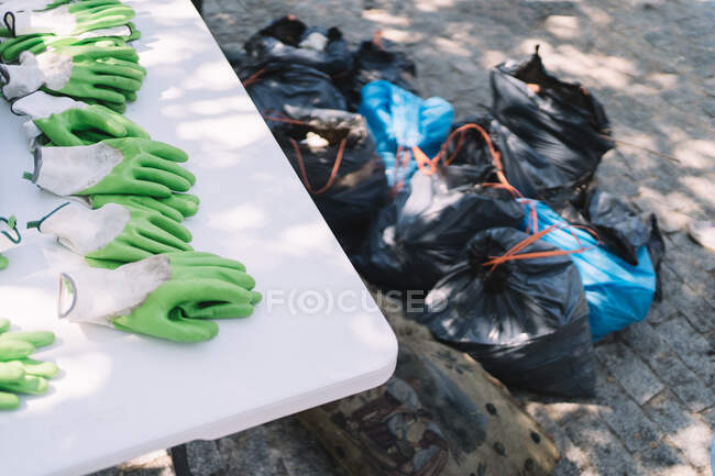 Gants en caoutchouc vert placés sur la table près du tas de sacs poubelle pendant la campagne de nettoyage de l'environnement dans le parc d'été — Photo de stock