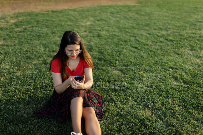 Женщина в летней одежде, сидящая на зеленом лугу в парке и серфинг Интернет на мобильном телефоне во время развлечений в выходные дни вечером — стоковое фото