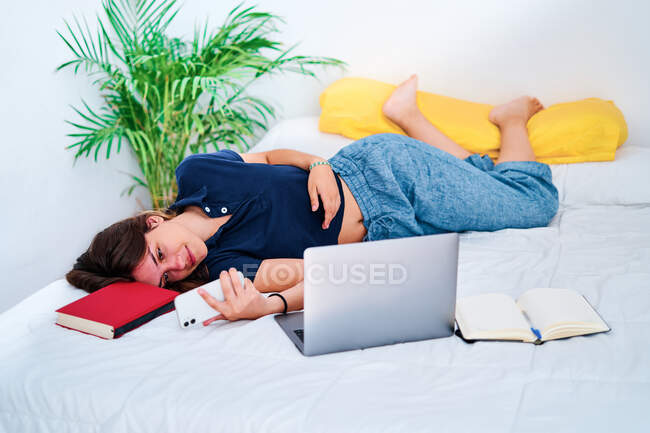 Молодая студентка лежит в постели с ноутбуком и учебниками и отправляет сообщения на смартфон во время дистанционного онлайн-обучения дома — стоковое фото