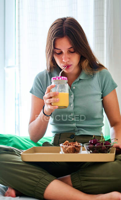 Молода жінка насолоджується здоровим сніданком зі свіжими ягодами та соком, сидячи з ногами, схрещеними на ліжку вранці — стокове фото
