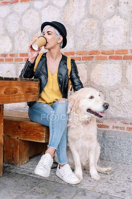 Full body of stylish young female owner in trendy outfit and hat drinking takeaway coffee while sitting on wooden bench on street with adorable Golden Retriever dog — Stock Photo
