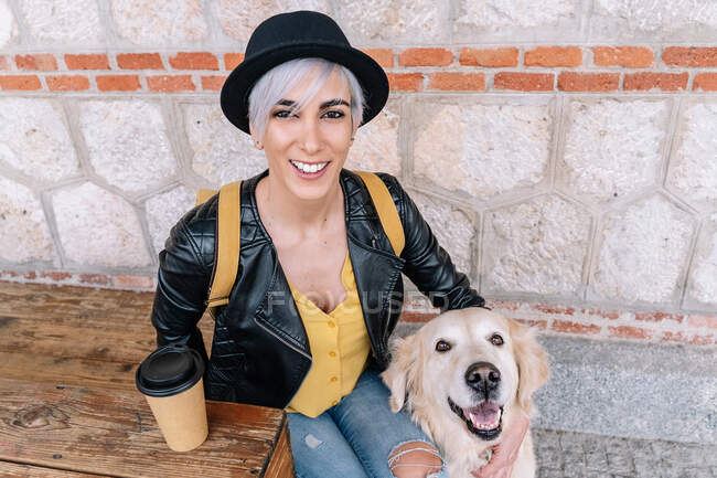 From above happy stylish young female owner in trendy outfit and hat drinking takeaway coffee while sitting on wooden bench on street with adorable Golden Retriever dog — Stock Photo