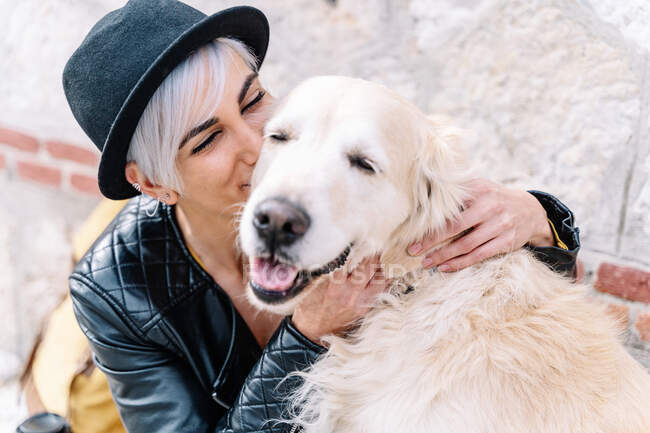 Trendy young female owner with dyed hair in stylish leather jacket and hat hugging and kissing adorable Golden Retriever dog while sitting on stone bench on street — Stock Photo